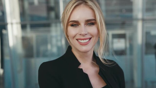Attractive young blonde woman in a stylish black outfit looks right towards the camera and smiles brightly. Airport terminal (business centre) on the background. Natural beauty, successful lifestyle.