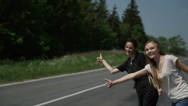 Two young women hitchhikers stand on the sidelines and ask to stop the passing cars