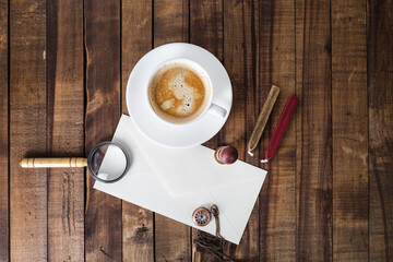 Blank envelope, magnifier, seal, stamp, clock and coffee cup on vintage wooden table background. Still life with postal accessories. Responsive design mockup.