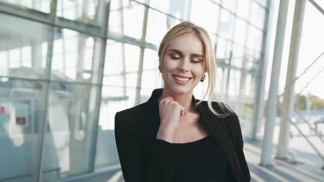 Gorgeous blonde woman in a black outfit looks right towards the camera and shares a beautiful smile. Airport terminal (business centre) on the background. Female portrait, cheerful mood. Modern woman.