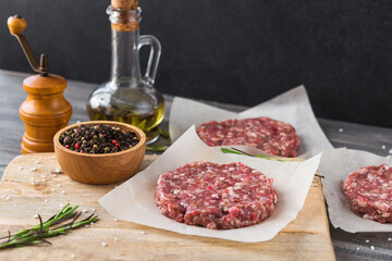 On a wooden cutting board on kraft paper there are raw beef burgers for burgers, a bottle of olive oil, spices, rosemary, a pepper mill, a cooking process, a top view