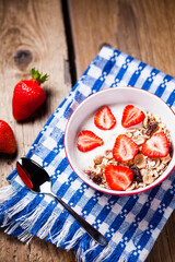 Granola with yogurt and strawberries in a red bowl on blue tablecloth on a wooden table. Red strawberries.