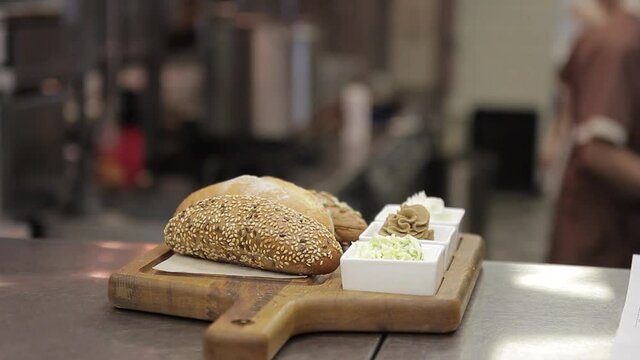 On a wooden tray lie buns with sesame and plates with condiments. In a large restaurant kitchen is ready to take out a plate with bread and different sauces and mustard. In the background, employees