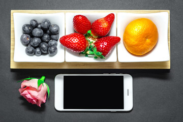 Zestaw owoców takich jak jagody truskawki mandarynka w białych pojemniczkach na czarne ciemne tło i biały telefon z czarnym pustym ekranem