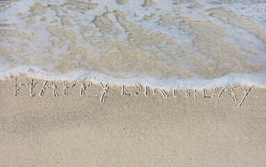 happy birthday written in beach sand with frothy ocean water
