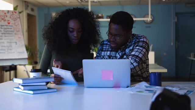 A young African American man with glasses and beautiful young African American woman with curly hair talking about business plans and working on a laptop in the office at night