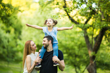 Father, mother and young daughter on piggyback with raised hands like flying on summer park background