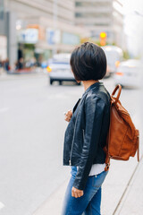 Obraz premium Portrait of young girl woman with short dark hair bob style, in blue jeans and leather biker jacket, with yellow brown backpack on shoulder, standing in city street, view from back behind