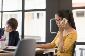 Charming casual business asian woman using smartphone and note something on book in modern office team meeting background.