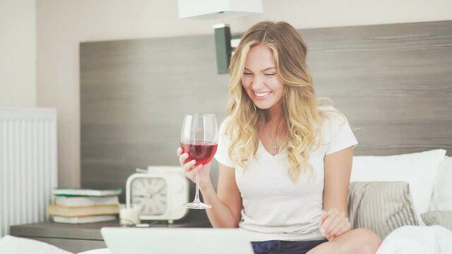 A woman smiles beautifully, drinks wine and communicates on the Internet using a laptop. Beautiful young woman in her bright bedroom drinking wine and talking on the Internet using a laptop.