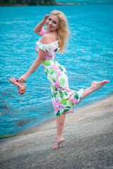 Beautiful blonde hair woman in romantic flowers dress near blue water and sand. Cute happy girl have fun on a nature