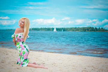 Fototapeta premium Beautiful blonde hair woman in romantic flowers dress near blue water and sand. Cute happy girl have fun on a nature
