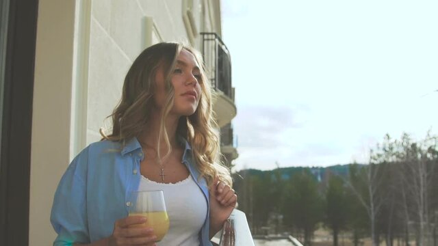 Woman on the balcony in the early morning and drinks orange juice. Attractive blonde drinking fresh on terrace. A romantic girl is standing on the veranda, looking into the distance and drinking juice