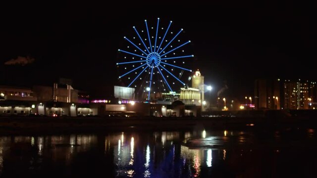 Time lapse of beautiful Ferris wheel illuminated with colorful lights spinning in the night in city center. View from river embankment