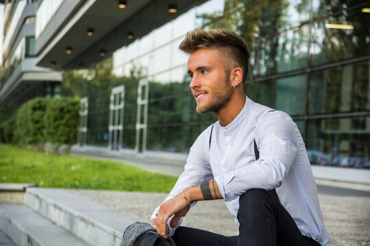 Stylish trendy young man sitting outdoor against office window, looking confident away