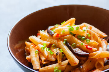 Close up of italian penne pasta with eggplants and tomato sauce in a brown bowl on a white stone background. Copy space and overhead view. Traditional food concept.