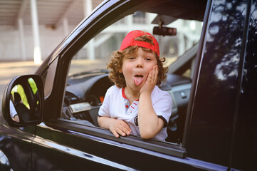 child with curly hair and a red cap sits behind the wheel of a car. baby boy grimaces in car window