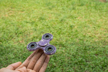 A hand holding fidget spinner purple colour spinning stress relieving toy on green grass background.