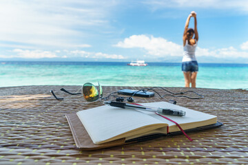 Opened empty notepad is on the table with sunglasses, phone, headphones at the background of tropical sea and relaxing girl