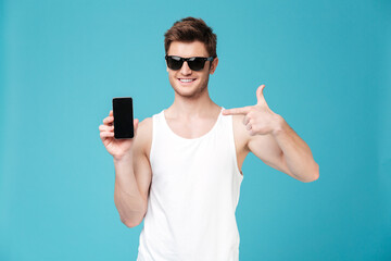 Young man pointing at blank screen of smartphone isolated over blue