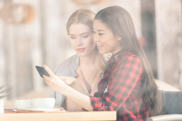 Smiling young women using smartphone while drinking coffee together at lunch meeting