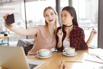 Smiling young women taking selfie while drinking coffee indoors at lunch meeting