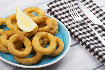 Close-up of platter of squids a la romana and a lemon on white wooden table next to a fork and a knife. Squids.