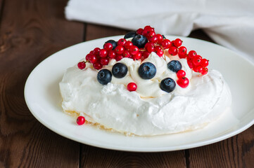 Close up of homemade meringue cake Pavlova with whipped cream with mascarpone, fresh blueberries and red currants in a white plate on a wooden background.