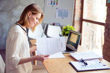Cute young blond lady is concentreted on ideas for new start up. She is in casual shirt, sitting at her work place and studying documents
