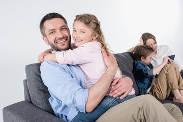 father with daughter embracing and looking at camera while mother with son sitting at sofa