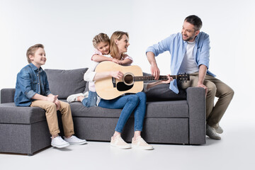 Happy young family with two children sitting together on couch and playing guitar