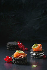 Stack of black wholegrain charcoal crackers with smoked salmon, cream cheese, green salad and red currant berries over black metal background. Appetizer snack. Space for text