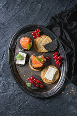 Stack of black charcoal and traditional crackers with smoked salmon, cream cheese, green salad and red currant berries on vintage metal tray over black stone background. Appetizer snack. Top view
