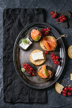Stack of black charcoal and traditional crackers with smoked salmon, cream cheese, green salad and red currant berries on vintage metal tray over black stone background. Appetizer snack. Top view