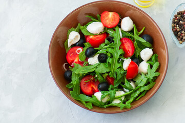 Close up of fresh salad with rucola, tomatoes  olives, onion, mozzarella and spices in a bowl on a white stone background with copy space. Healthy food, Diet, Detox, Clean Eating or Vegetarian concept