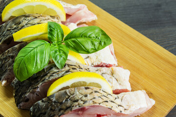 Sliced pieces of raw fish on a wooden board with lemon and basil leaves prepared for baking. Selective focus.