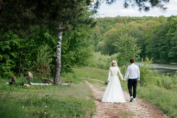 Beautiful bride and groom go holding hands in the wood near river, back view