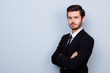 Young confident man in black suit with crossed hands standing half-turned against gray baclground