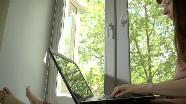 Young attractive woman in beige overalls using laptop sitting on window sill in sunny spring day outdoors green trees
