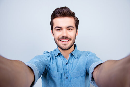 Handsome brunet young man is making selfie and smiling. He is wearing jeans shirt and behind him is a light blue background