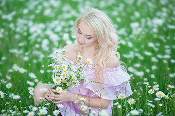 Attractive blonde in chamomile field. Young woman in wreath surrounded by chamomiles wearing stylish pink dress enjoying summer spring
