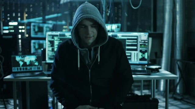 Dangerous Internationally Wanted  Hacker Speaks into the Camera. In the Background His Operating Room with Multiple Displays and Cables. Shot on RED EPIC-W 8K Helium Cinema Camera.