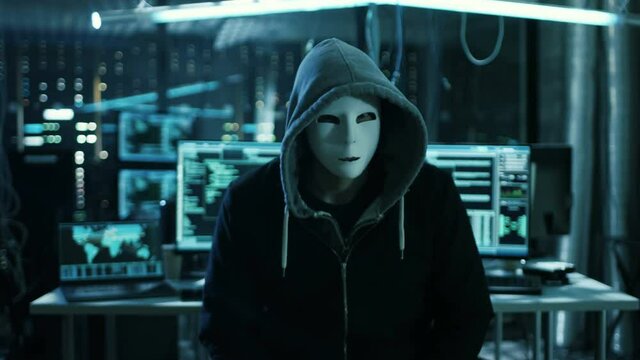 Dangerous Internationally Wanted Hacker in a Mask Speaks into the Camera. In the Background His Dark Operating Room with Multiple Displays and Cables.Shot on RED EPIC-W 8K Helium Cinema Camera.