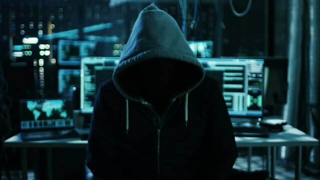 Dangerous Internationally Wanted Hacker with Covered Face Speaks into the Camera. In the Background His Operating Room with Multiple Displays and Cables. Shot on RED EPIC-W 8K Helium Cinema Camera.