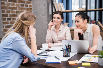 beautiful smiling multicultural businesswomen discussing project at workplace with laptop