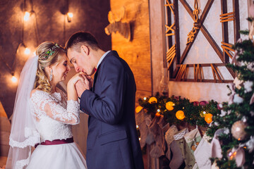 Bride in beautiful dress and groom in gray suit sitting on sofa indoors in white studio interior like at home. Newlyweds kiss each other at home. Flowers bouquet in woman hand.