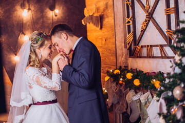 Bride in beautiful dress and groom in gray suit sitting on sofa indoors in white studio interior like at home. Newlyweds kiss each other at home. Flowers bouquet in woman hand.