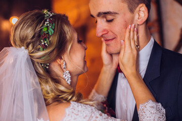 Bride in beautiful dress and groom in gray suit sitting on sofa indoors in white studio interior like at home. Newlyweds kiss each other at home. Flowers bouquet in woman hand.