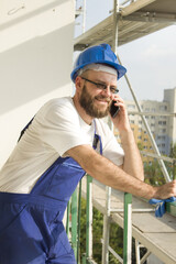 Uśmiechnięty pracownik budowlany w stroju roboczym i w kasku na głowie rozmawia przez telefon. Praca na dużej wysokości. Rusztowania w tle. - obrazy, fototapety, plakaty