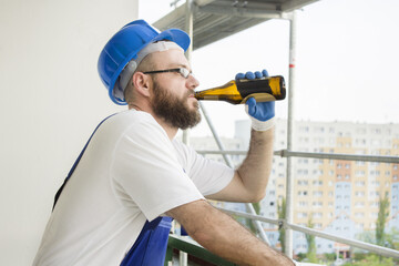 Pracownik budowlany w stroju roboczym, rękawicach ochronnych i kasku na głowie pije piwo z butelki. Praca na dużej wysokości. Rusztowania w tle.